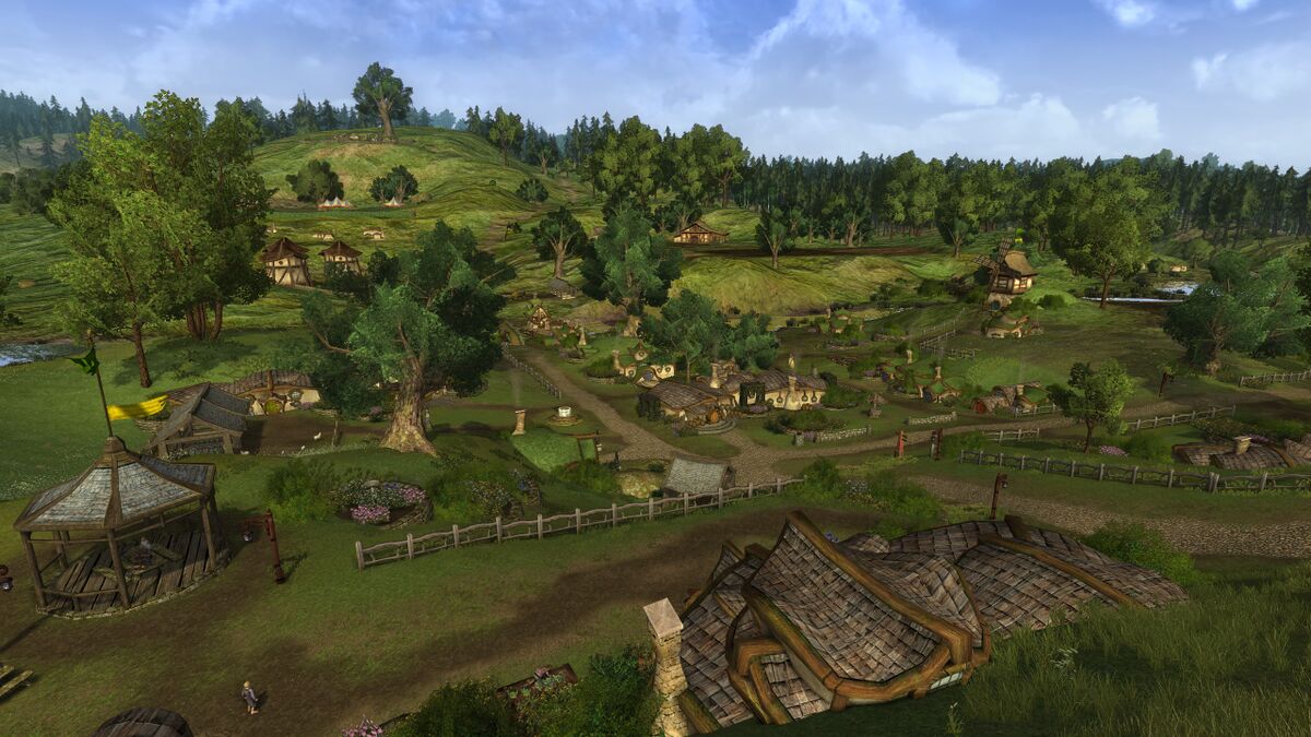 A daytime view over Hobbiton in LOTRO, with branching roads, fenced gardens, hobbit-holes and mature trees beneath a blue sky.